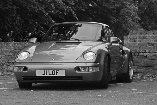 A black and white photo of a parked car - Pistonheads - The image is a black and white photograph of a parked sports car. The car has a clean, sleek design and features a silver theme with the tagline "JI LOF" on the license plate. The vehicle is positioned in front of a brick wall and appears to be on a street mainly characterized by trees in the background.