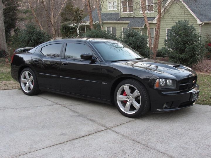 Possibly Buying a Charger Hemi SRT8..  WTF..? Opinions pls - Page 1 - Yank Motors - PistonHeads - The black Dodge Challenger sits on a concrete driveway, giving off an air of power and elegance. Its large silver wheels and shiny black exterior reflect the surrounding neighborhood of green-colored houses and trees. The car is positioned facing towards the right side of the image, ready to confidently navigate the suburban streets. The daylight suggests it's an ideal time for a drive in this beautiful borough.