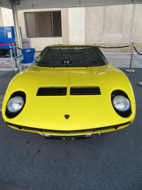 Pistonheads Classic Emaar - The image features a bright yellow sports car, parked on a tiled surface with a meta grill design noticeable on the front. The vehicle is located indoors, under a covered area, possibly a show-car display space. To the left, there's a blue chair, and nearby, a number "13" is visible, possibly indicating the car's exhibition number or some sort of serial identification. The background is not clearly visible due to the focus on the sports car.