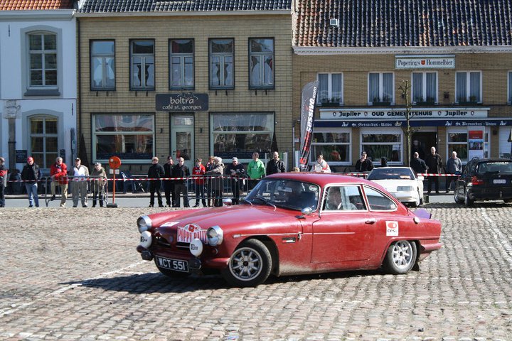 Minilite wheels love -in. - Page 1 - Classic Cars and Yesterday's Heroes - PistonHeads - The image shows a vintage, red, two-door sedan parked on a stone-paved street. In front of the car, a small crowd is observing the car. The street is lined with storefronts to both sides, and there's an event setup with banners. The overall scene suggests a casual gathering or perhaps a mini antique car show.