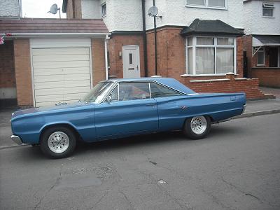 71 GTO in classifieds, what has it not sold? - Page 1 - Yank Motors - PistonHeads - The image depicts a classic blue car parked outside a two-story brick house with a large white garage. The garage door is closed, and there seems to be another vehicle visible inside the garage. The car has white-walled rims and appears to be an older model, potentially from the 1960s or 1970s. A sidewalk is present next to the parked car, and the street looks quiet with no other traffic visible.