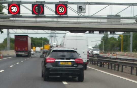 A car driving down a street next to a traffic light - Pistonheads - The image captures a lively traffic scene on a highway. The photo is taken from the rear of a vehicle, following a line of cars. Traffic signs reading "50" and "50" are visible, indicating a 50 km/h speed limit. Various vehicles, including cars, a truck, and a bus, are present, all moving in the same direction. The bridge above the vehicles is part of an infrastructure system.