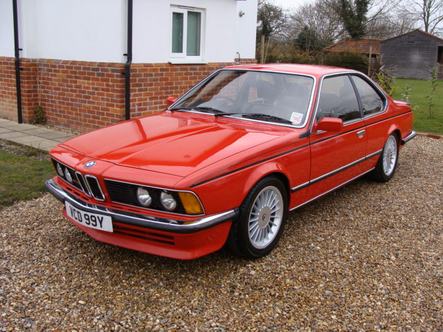 Old newbie - Page 1 - Readers' Cars - PistonHeads - This image features a vibrant red BMW parked on a gravel driveway. The BMW is facing the camera, sharply angled slightly to the left, showcasing its prominent front grille and sleek design. The car's reg number is clearly visible as "VCD 99Y". In the background, a white building can be seen, partially obscured by the car, and some green vegetation in front of the building. The overall scene gives a sense of a quiet residential area.