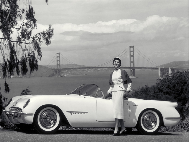 Corvette Evolution: How it Became an American Legend - Page 1 - Corvettes - PistonHeads - In the sepia-toned black and white image, a woman exudes elegance, standing beside a vintage white convertible car. The car is parked by a coastal road that offers a view of the Golden Gate Bridge in the background. Overhanging trees add to the serene and picturesque scene. The woman's attire, detailed with a cardigan, suggests a touch of vintage charm complementing the age of the vehicle. This image seems to capture a nostalgic moment, perhaps a travelogue of a journey down the California coast.