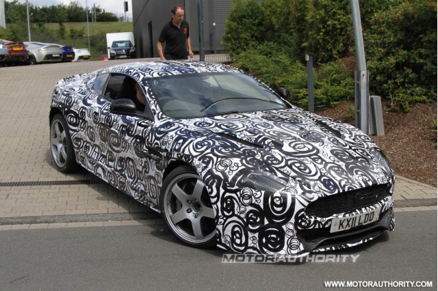 New DB9 /DBS? - Page 1 - Aston Martin - PistonHeads - This image showcases a black sports car with a unique, intricate camouflage pattern that covers the entire vehicle. The design of the camouflage consists of continuous, flowing lines that cross over each other, creating a complex, geometric appearance. The man standing behind the car seems to be inspecting or admiring it, and the car is parked on a street next to a building. There are also some other cars visible in the background, parked at various distances from the foreground car.