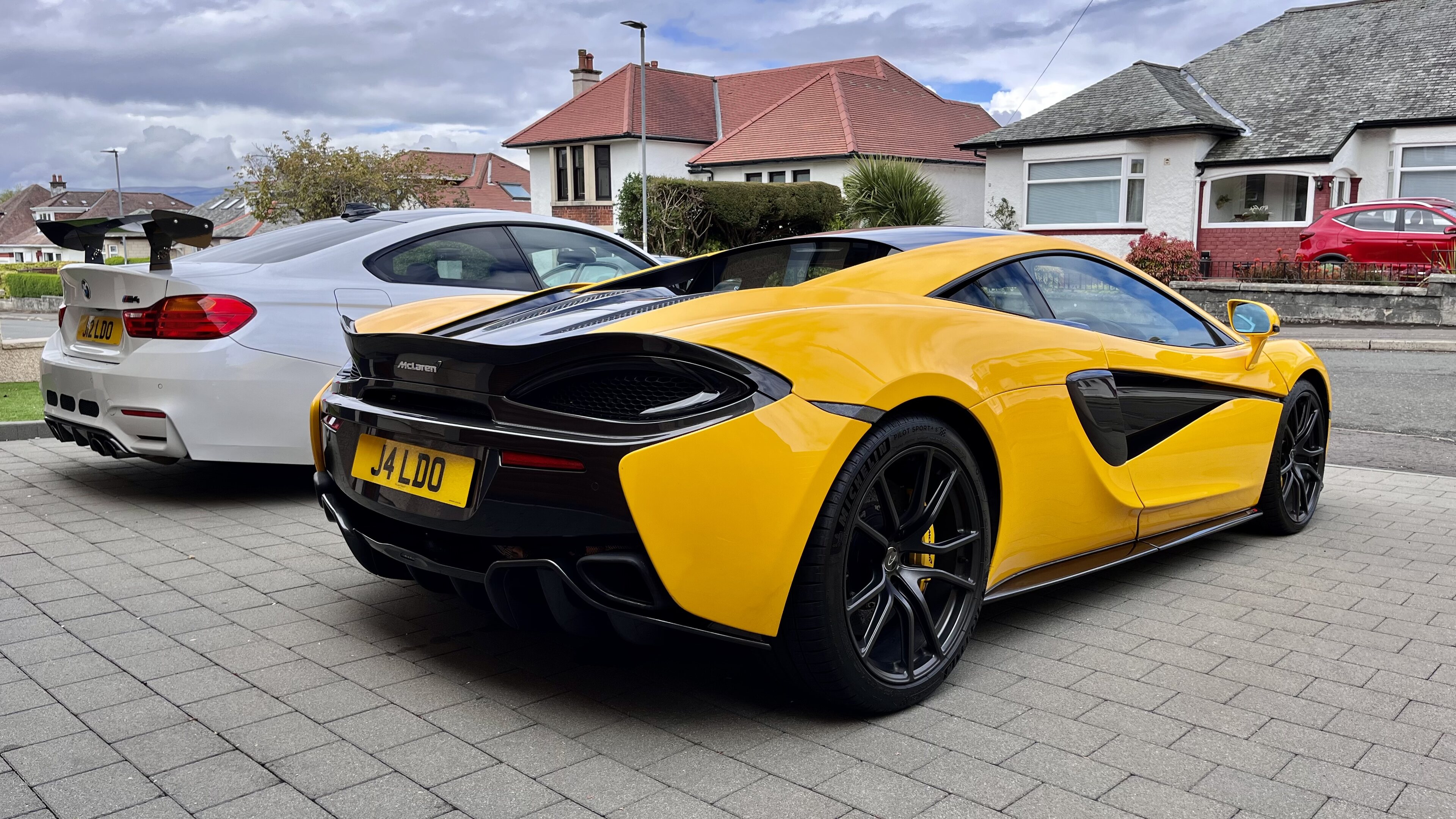 Volcano Yellow 570s - Page 9 - McLaren - PistonHeads UK - The image shows a vibrant yellow Aston Martin sports car parked on the street. It appears to be an Aston Martin DB11, characterized by its sleek lines and distinctive design elements. The car is positioned in front of two other vehicles, one white sedan and another, partially visible, black SUV. The setting suggests an urban environment with residential buildings lining the street, providing a backdrop that contrasts with the luxury vehicle.