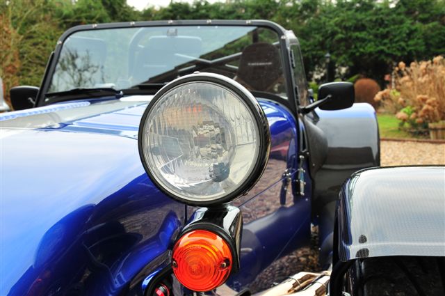 Any Seven owners in Barcelona? - Page 1 - Caterham - PistonHeads - The image showcases a bright blue convertible car parked outdoors. The car's hood and roof are a darker shade of blue, lending a sporty contrast to the car's body. The front part of the car is prominently featured, with a clear view of the headlight and orange turn signal indicators. The car is parked in a location where trees can be seen in the background, suggesting a residential street or a parking area near a park or forested zone. The overall lighting and composition of the image suggest it was taken during the day with pleasant weather.