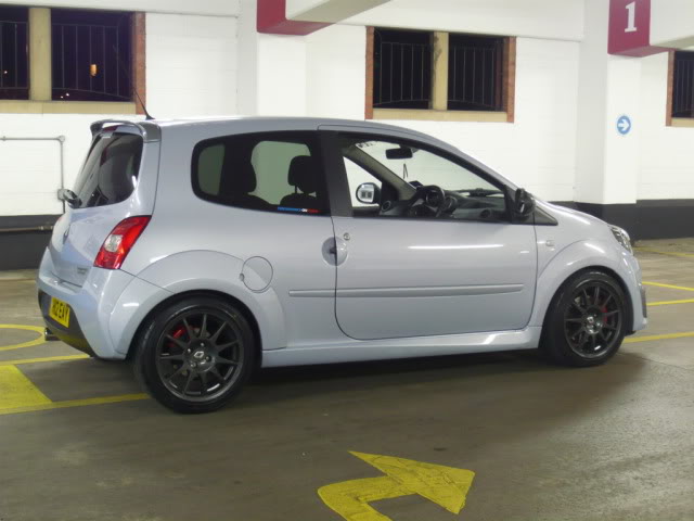 Twingo Newbie Owner Pistonheads - The image shows a small hatchback car parked in a covered parking area. The car is silver in color, with a black roof rack and black wheel rims. There is a yellow arrow on the ground, indicating a turn or maneuver. The floor area where the car is parked seems to be bordered by yellow lines, possibly for reserved spaces or parking restrictions. The car appears to be in good condition, without any visible damage or paint issues.