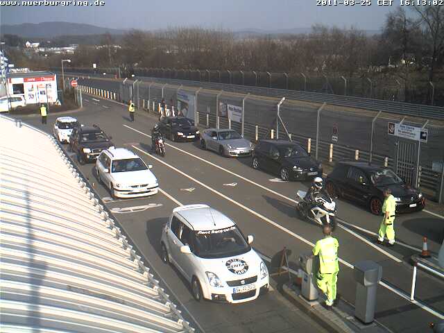 Webcam Ring Pistonheads - The image shows a bustling scene with several vehicles stopped in a line. There are multiple cars, a truck, and a motorcycle among them. Some individuals are present with one person on a motorcycle and another near the vehicles. The environment suggests it might be a special event or a busy traffic situation as evidenced by the differing directions of the vehicles. The place looks outdoors, and there are fences and a sloped roof in the backdrop, suggesting a parking lot or a similar structure.