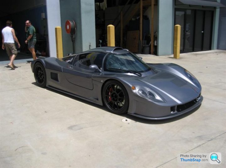 Pistonheads - The image presents a sleek, silver racing car, parked in a lot. It's parked next to a garage, its polished surface reflecting the surroundings. The car appears to be parked outdoors, as indicated by the brightness of the surroundings. In the background, there are a few people, possibly related to the car, as they seem to be in the vicinity of the car and an airplane. The presence of the airplane suggests that the location could be an airport or an aviation-related facility.