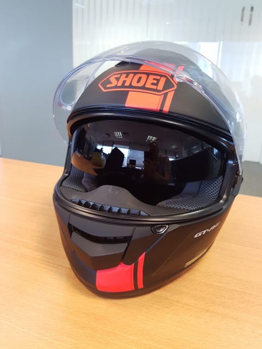 A close up of a baseball glove on a table - The image features a black and red helmet with the "Shoei" brand logo prominently displayed at the top of the helmet's peak. The helmet appears to be made of a plastic or polycarbonate material, typical for motorcycle riding helmets. The interior of the helmet is visible through the front visor and is not stereotypically designed to resemble a spacecraft. The helmet is placed on a wooden surface, which provides a natural, neutral background that contrasts with the helmet's sleek, modern design. The left side of the helmet (from the viewer's perspective) is facing the camera, providing a clear view of the logo and the design details that are indicative of the brand.