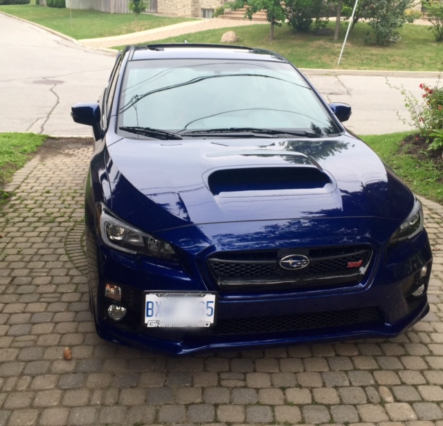 A car parked next to a parking meter - Pistonheads - The image shows a dark-colored sports car parked on a driveway in front of a house. The car's sleek design is highlighted by its glossy finish, and it prominently displays a license plate on the front. The driveway appears to be made of bricks, and there is greenery and a grassy area visible in the background. There's a slight reflection of the car on the surface it is parked on, indicating it might be a sunny day. There is also a small object lying near the front tire of the car.