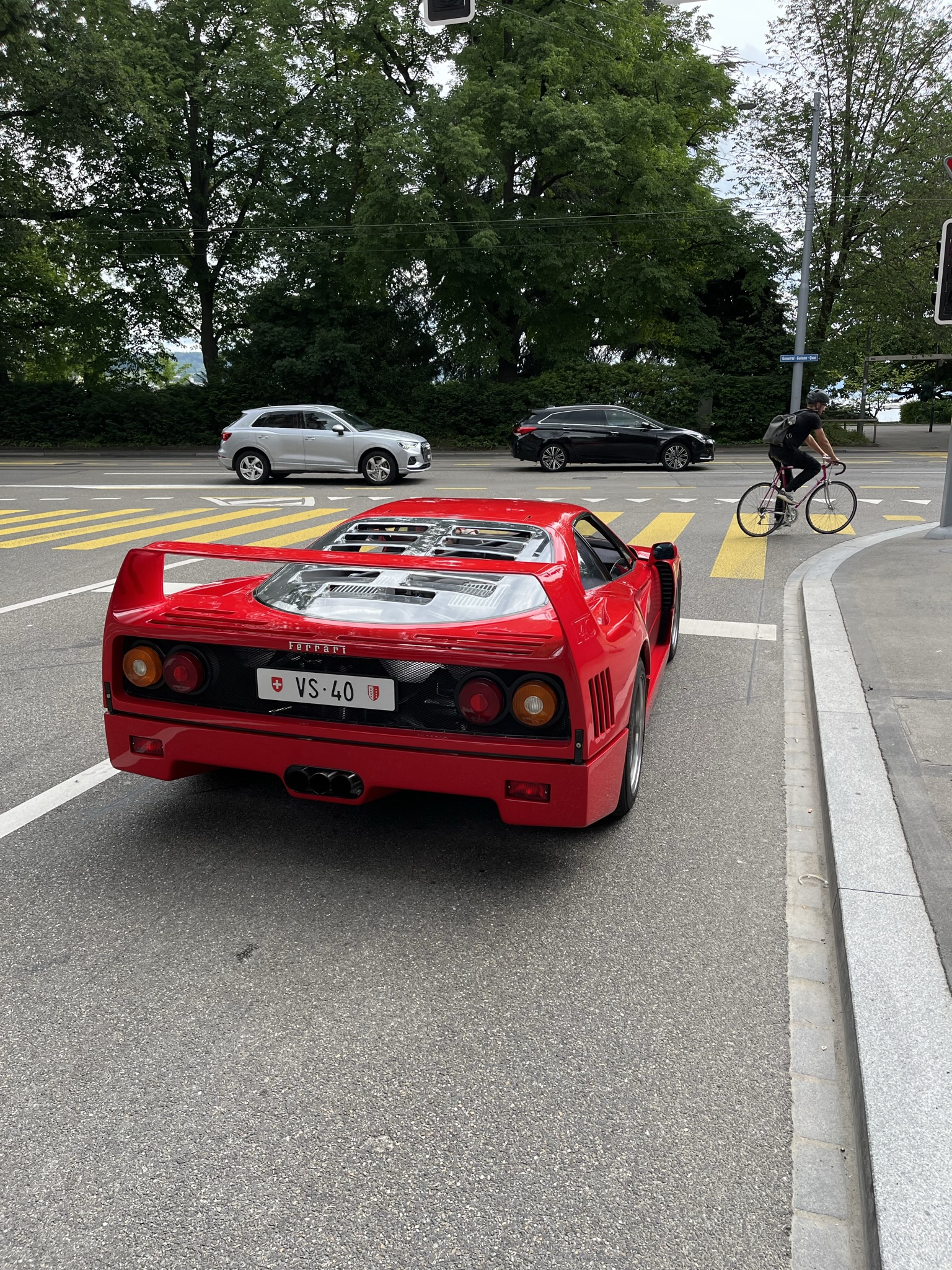 Supercars spotted, some rarities (vol 7) - Page 1 - General Gassing - PistonHeads UK - The image depicts a vibrant red Ferrari sports car parked on the side of a road. The Ferrari is prominently positioned, taking up a large portion of the frame. It features a license plate indicating that it's from Italy. In the background, there are cars and pedestrians going about their day, suggesting an urban setting. A traffic light is also visible, reinforcing the street-side location. The car itself seems to be well-maintained, with no visible damage or wear.