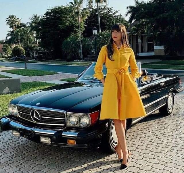 Best smoker barges 1-5 large [Vol 19] - Page 169 - General Gassing - PistonHeads UK - This is a color photograph featuring a woman standing next to a vintage Mercedes-Benz car. The car, with its distinctive hood ornament and classic design, is black in color. The woman is dressed in a vibrant yellow coat that contrasts with the darker tones of her surroundings, including the paved surface of what appears to be a residential area with palm trees in the background. She is also wearing a dark skirt, high heels, and has long, flowing hair. Her attire suggests a formal or special occasion, and she stands confidently beside the car, which adds an air of elegance to the scene. The image exudes a sense of style and sophistication associated with luxury vehicles and upscale living.