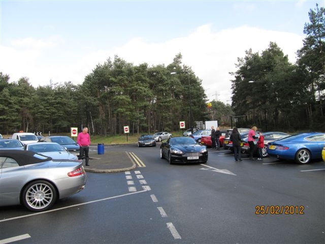 Fleet Services this morning - Page 1 - Aston Martin - PistonHeads - The image portrays a bustling scene at an outdoor parking lot, filled with parked cars, with a few people scattered around. The lot is situated amidst a lush green forest, with trees standing tall in the background. Amidst the vehicles of various colors and models, there are a few distinct signs, including one with a red circle prominently displayed. The sky above is clear, suggesting it might be a warm, sunny day. From the timestamp in the corner, the photo was likely taken on March 24, 2012.