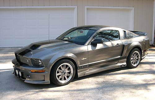 Pistonheads Mustang Buy - The image shows a silver Mustang parked on a concrete driveway. The car is in good condition, with a shiny exterior that reflects the light. It features a convertible top, which is closed, revealing tinted windows and a visible back window. The vehicle is equipped with five-spoke alloy wheels. To the right of the car, there is a garage door partially visible with a white panel and a door entrance screen. The driveway appears to have slight shadowing, indicating that there is some sunlight. The image appears to be a photograph taken during daytime.