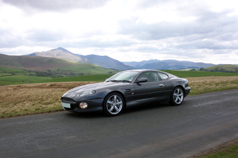 Free Introduction Pistonheads Photoshoot - This image shows a curvy black sports car driving on a paved road, with a backdrop of mountainous terrain. The day is partly cloudy, suggesting it could be either morning or afternoon. The car's taillights are visible, and there is a grassy embarkment to the side of the road. The vehicle appears to be in motion, suggesting speed or travel. The overall scene conveys a sense of freedom and adventure.