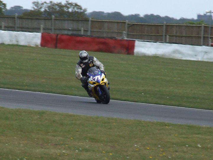 Front Pistonheads Losing Afraid - The image captures a thrilling moment on a race track. A person, clad in a black jacket and a yellow helmet, is skillfully riding a motorcycle. The motorcycle and rider are highlighted against the race track, which contrasts sharply with the surrounding grass. Subtle hints of caution, like a red and white barrier, remind us of the high-speed nature of the scene. The rider is making a turn on the track, maintaining a sleek racing posture.