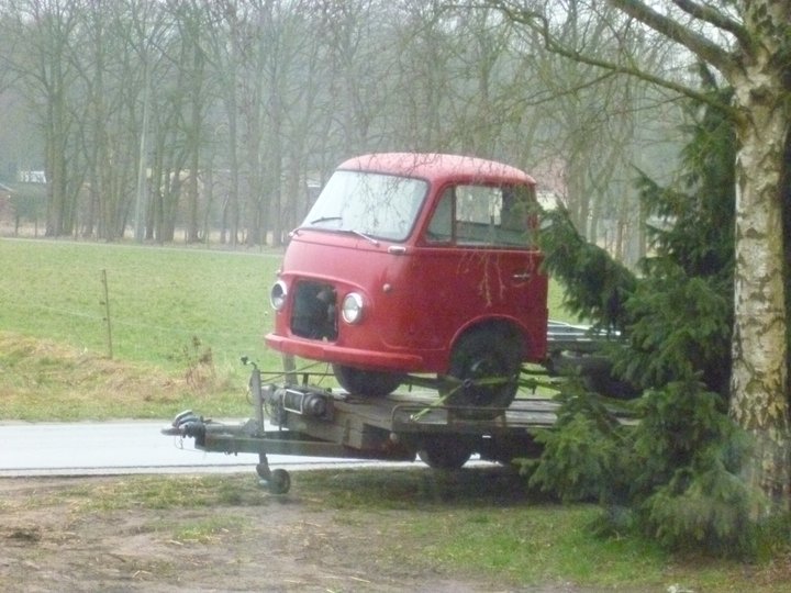 Taunus Pistonheads Transit - The image captures a rusted red vintage truck, which appears to be an antique wagon, cleverly transported on a trailer by a gray modern car. The truck is prominently featured centered on a large trailer, creating a striking contrast between the old and the new. The car and trailer combination suggests a careful maneuver take place on an overcast day, possibly hinting at an impending rain or perhaps showcasing an event or curiosity towards classic vehicles. The background reveals a field and a row of trees, adding to the rural and peaceful setting of the scene.