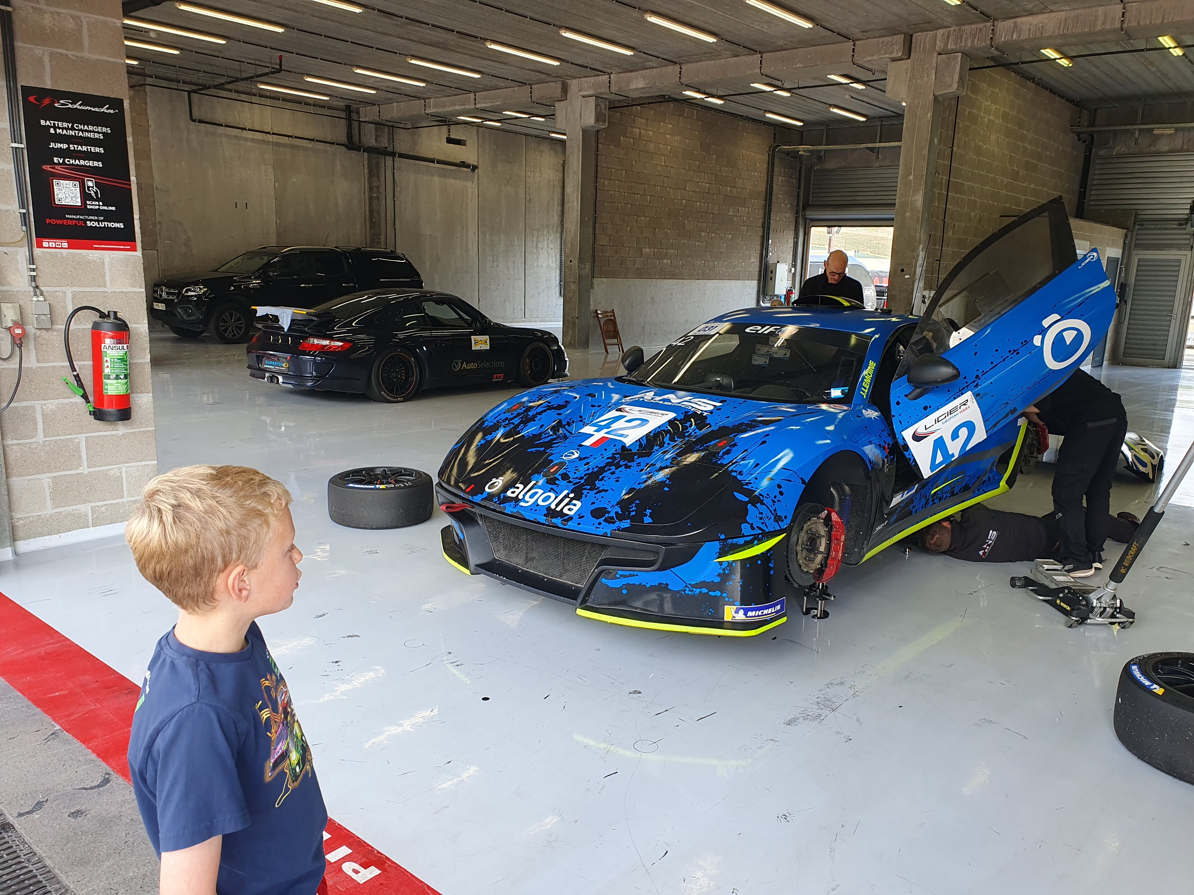 Pistonheads - In the image, a young boy stands in front of a blue and white Formula 1 race car. The car is parked inside a garage filled with various other cars, suggesting a racing or automotive event. The child appears curious and interested in the car, looking towards it with his hands tucked into his pockets. A man, possibly an employee or a family member, stands to the side of the boy, watching the interaction between the child and the race car. The setting is industrial, characterized by concrete floors and metal walls typical of garages, emphasizing the mechanical nature of the scene.