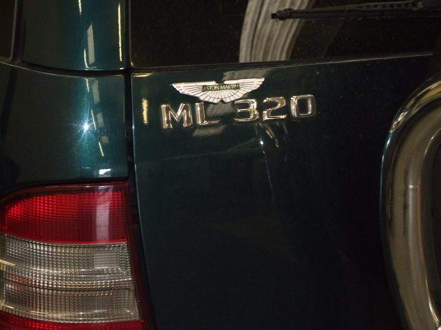 Light Pistonheads Shed - The image shows a close-up of a car's rear side door, which features a logo. The logo reads "ML320," suggesting that this is a luxury vehicle. The design of the car is dark, with a visible antenna and a silver brake light cover. The car is parked in a garage or industrial setting, as indicated by the presence of a concrete wall and mechanical parts in the background.