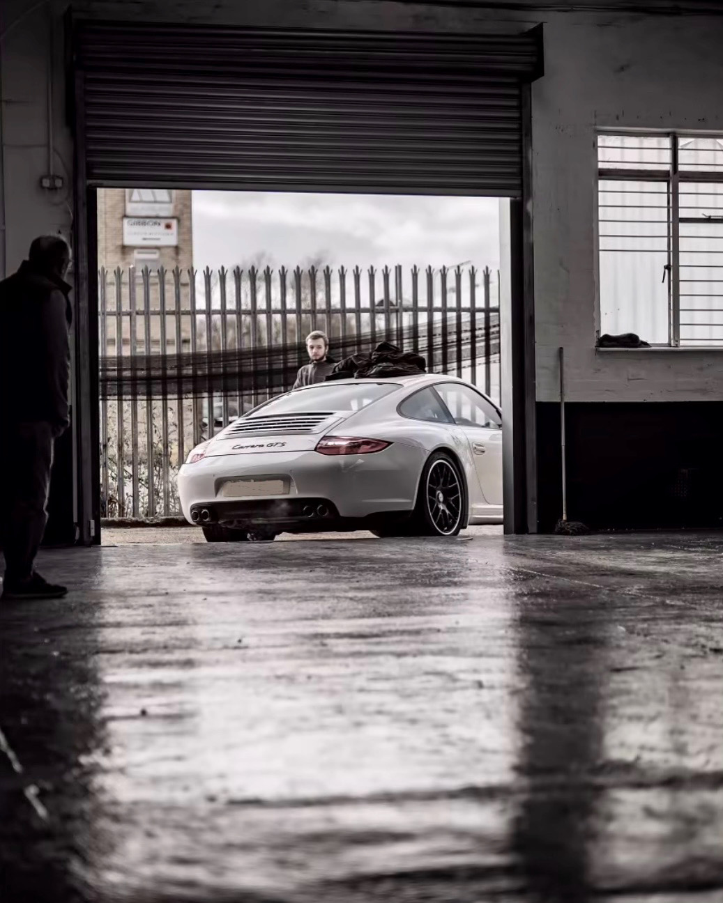 Pistonheads - This is a black and white photo of an indoor setting, featuring a large sports car parked inside a garage. The vehicle, with its sleek design and shiny surface, takes up a significant portion of the space and stands out prominently against the darker background. There are two individuals present in the scene; one person is partially visible near the car, while another individual is positioned farther away, standing close to a metal door. The lighting within the garage casts shadows on the floor, enhancing the contrast between light and shadow in this photograph. The overall atmosphere of the image conveys a sense of solitude and contemplation.