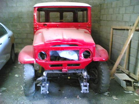 Toyota FJ40 - Page 1 - Off Road - PistonHeads - In the image, a large, red vintage truck is the main object, parked in a concrete enclosed area with walls visible. The truck appears to be in the process of being restored, evident by the scrap materials scattered around it. It has massive wheels and has been repainted, but some parts like the hood are showing signs of wear. There's no visible driver inside the vehicle.