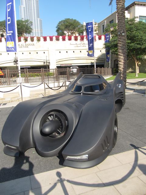 Pistonheads Classic Emaar - The image showcases a formidable black Batmobile replica parked on a concrete sidewalk. This large scale replica boasts a sleek design with a distinct curved front, which adds to its futuristic and intimidating appearance. The side of the car features a large exhaust, adding to the emphasis on its powerful nature. The car is displaying a single headlight on the passenger side, an iconic feature of the Batmobile. In the background, there are palm trees lining the street, providing a contrasting environment to the showcased car. The building behind the car has several flags and banners, hinting at an urban setting possibly in a city center or a popular tourist area.