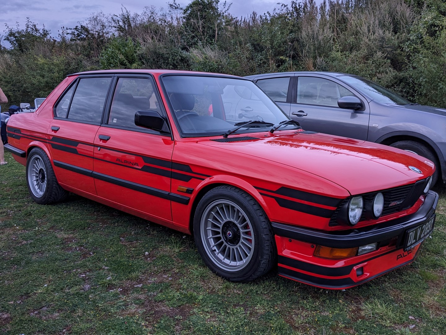 M3 CSL and M5 - Page 9 - Readers' Cars - PistonHeads UK - The image shows a red and black vintage car parked on grass. The vehicle has distinctive stripes running along its body and a large rear spoiler. It's a sunny day, with the sky visible in the background. There is a person standing behind the car, partially obscured by it. In the distance, there are other cars that are less prominent than the main red car. The setting appears to be an outdoor event or exhibition.