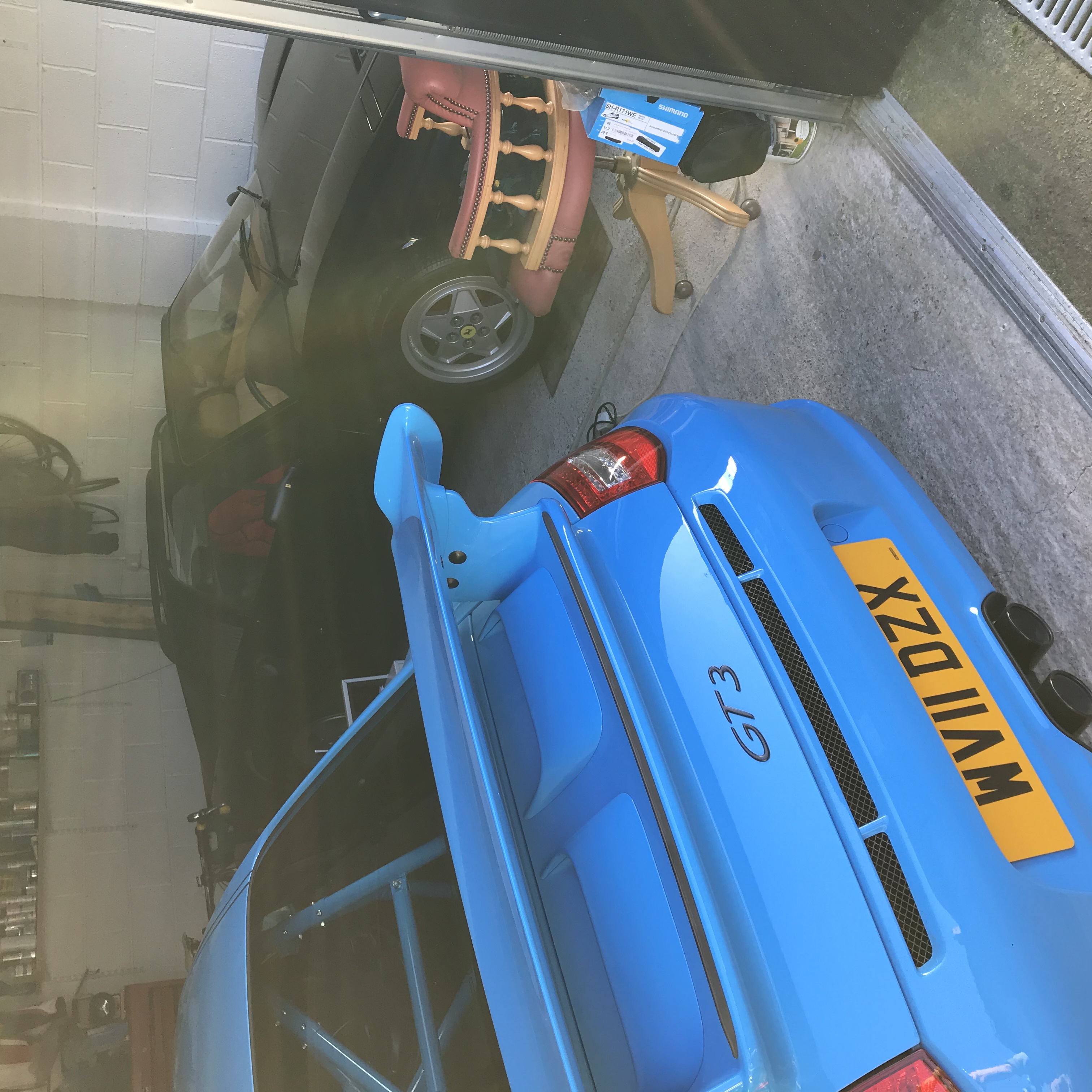 Pistonheads - The image shows a garage interior where two cars are parked, one blue and the other blue with white stripes. There are also some personal items visible, such as a chair, a couch, and a television. A small portion of a dining table can be seen in the background. Natural light is coming through the windows, suggesting it's daytime. The garage appears to be well-maintained and organized.