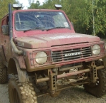 Santana Pistonheads Newbie - The image showcases an off-road vehicle, possibly a jeep or a pickup truck, which is heavily covered in mud. The vehicle is predominantly red, with the front and roof visibly dirtier due to the environmental conditions. The side profile of the vehicle reveals a robust and rugged design, typical for off-road models, and there are no visible texts or branding. The background is dimly lit, with the foreground vehicle occupying most of the image's space, which suggests that the focus is on the vehicle itself rather than its surroundings.