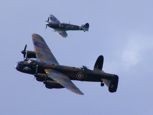 Pistonheads - This image captures a dramatic scene of two military aircraft in mid-flight, flying side by side in the clear blue sky. The aircraft of the foreground is a dark brown bomber with white markings, bearing the letters "HMR" prominently on its side. The other aircraft, slightly higher in the sky, is an older style fighter plane, decorated with circular and cross-like designs on the wings and fuselage. Both planes are generating power, as evidenced by the propeller visible on the bomber. The clear sky and lack of contrails suggest high altitude and possibly the beginning or end of a flight mission.