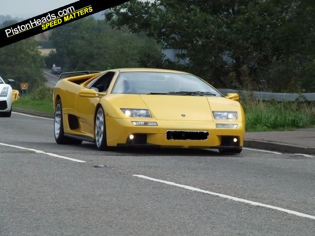 Pistonheads Supercar - The image shows a bright yellow convertible sports car in motion on a road. The car is distinctive with its large, skinny side mirrors and prominent front bumper. The road itself has white lane markings suggesting it is a two-lane road with lanes designated by a double dashed line. To the side of the road, there is green vegetation and grass, indicating the presence of a grassy area or possibly a median strip separating the opposing lanes of the road. In the background, there are other vehicles visible at a considerable distance, suggesting that the car is part of a multi-lane road system or a highway. The overcast sky suggests cloudy weather conditions.