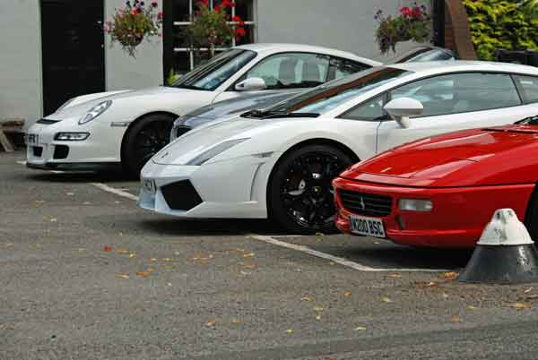 A car is parked on the side of the road - Pistonheads - The image captures a serene scene of a parking lot filled with three luxury cars parked side by side. The first car is a white Porsche, sleek and grand, showcasing its imposing presence. Adjacent to it, the second car is a vibrant red sports car, its shiny exterior reflecting the ambient light. The third car in the trio is a pristine white Porsche, mirroring the first one in design and color. 

The parking lot is not just home to these cars; it also houses several potted plants that add a touch of greenery to the urban landscape. A gray building forms the backdrop of this scene, providing an interesting contrast to the colorful cars. 

The cars are parked parallel to a striped curb, with the Porsches positioned closer to the building. The red sports car is parked slightly further away, creating a visually balanced composition. The overall scene exudes an air of elegance and sophistication.