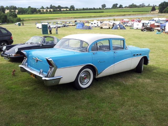 NSRA Hot Rod Supernats - Page 1 - Yank Motors - PistonHeads - The image shows a classic, old-fashioned car in a field, surrounded by green grass. The car is a vintage model, with a blue and white color scheme. In the background, there are several other cars and trucks of different makes and models. The setting appears to be an outdoor exhibition or event, with the car stationed in the center, drawing attention. The lighting suggests it might be early evening, creating soft shadows on the grassy ground.