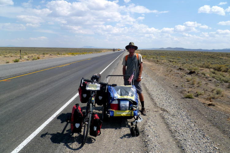 Lands end John o groats (walking) LEJOG - Page 27 - Holidays & Travel - PistonHeads UK - This is a color photograph showing an individual standing next to a motorcycle with luggage attached. The person is wearing a hat and appears to be preparing for travel, as suggested by the packed suitcases on the bike. They are outdoors in a dusty, arid environment, possibly on a road trip or an adventurous journey. In the background, there's a clear sky and a flat horizon that includes a stretch of highway with a single vehicle visible in the distance. The overall scene conveys a sense of anticipation for an upcoming journey or expedition.