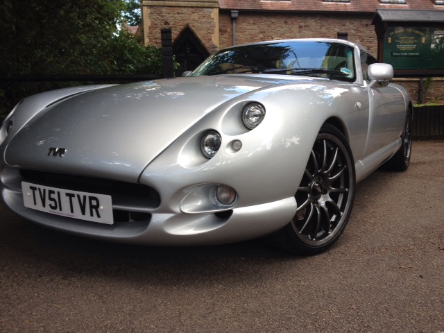 South West PH Car Photos inside... - Page 1 - South West - PistonHeads - The image shows a classic sports car, possibly a TVR, parked on a road by the side of a building. The car is silver with the model name startin with the letter "T". It features a distinctive, aerodynamic body with a long hood and a short rear deck. The car also has large, spoked alloy wheels and a prominent front grille. The license plate reads "TVS1TVR", and there's a registered trademark symbol indicating that "TVR" might be the brand of the car. In the background, there are trees, and behind the car, there's a stone building with a sign for "Tchuma".