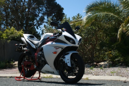 Pistonheads - The image showcases a white motorcycle parked on a street. The motorcycle features racing-style specs and is parked next to what appears to be a tropical environment, likely a palm tree. Attached to one of the motorcycle's wheels is a red kickstand, which is in use, supporting the motorcycle upright. The motorcycle is likely in a residential area, as there is a glimpse of a house in the frame but the focus is on the parked motorcycle.