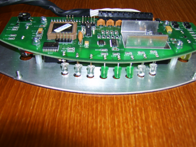 Pistonheads - The image depicts a close-up of a green circuit board lying flat on a wooden surface. The circuit board is populated with various electronic components, including resistors and capacitors, visible along the bottom and edges of the board. Multiple metallic pins extend from the board, suggesting it might interface with other components. On the right side of the board, there is a partially obscured connection, which might be a power cord or other type of interface. The focus is on the board, while the background is a blurred brown surface, indicating that the board is not secured or propped against anything else. The lighting seems to be artificial, casting shadows below the board and creating a somewhat textured appearance on the wooden surface.