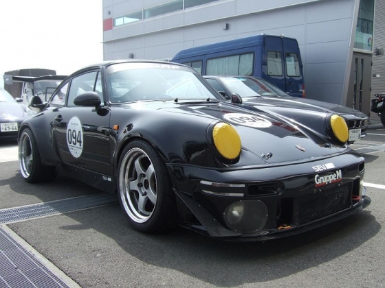 Better than a Speedline ? - Page 1 - Porsche Classics - PistonHeads - This image showcases a black Porsche car parked in front of a building. The Porsche is a sports car and has front and rear spoilers, distinctively orange clear headlights, and black and silver wheels. In the background, there are a few other cars parked, and the building appears to be a commercial structure with a formal facade. The Porsche is the main focus of the image, with its visible design and prominent placement.