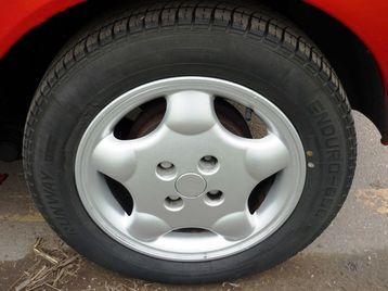 Pistonheads Megane Alloy Part - The image shows a close-up of a silver hubcap on a car's wheel. The hubcap has a sporty design with multiple rounded segments connected in a pattern. The wheel is not visible separately; the hubcap and part of the wheel cap extend outward from the center of the wheel, with the lug nuts visible in the middle. The image is a candid photograph taken on a road, possibly under some shade, as there's a subtle shadow under the hubcap. The focus is clear, highlighting the details of the hubcap design.