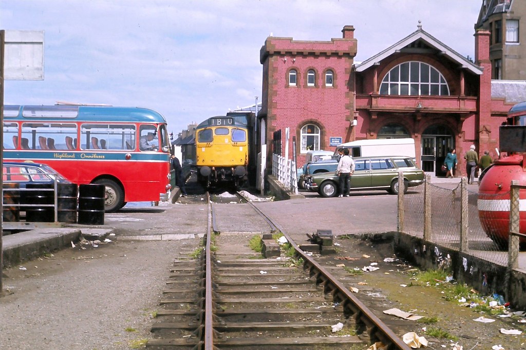 A 'period' classics pictures thread (Mk II) - Page 346 - Classic Cars and Yesterday's Heroes - PistonHeads UK - The image depicts a bustling train station with multiple trains present. A red bus is visible on the right side of the frame, suggesting a multimodal transportation hub. There are also several people scattered throughout the scene, likely passengers waiting for their respective trains or vehicles. The architecture of the train station suggests a historic setting with a brick structure and a clock tower in the background. The sky above appears overcast, casting a muted light over the scene.