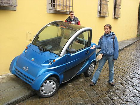 Insurance for a "Smart Labs", 'Open Smart' electric car - Page 1 - EV and Alternative Fuels - PistonHeads - The image features a blue smart car on a brick sidewalk. Two people are standing nearby. Both individuals are dressed in casual attire suitable for a cool day, one person is wearing a backpack, and they both appear to be smiling or laughing. The car's compact size and vibrant blue color make it stand out against the more subdued background of the brick pavement and nearby buildings. The people and the car suggest a light-hearted, possibly humorous, atmosphere. The identity of the people and the car model are not discernible from the image.