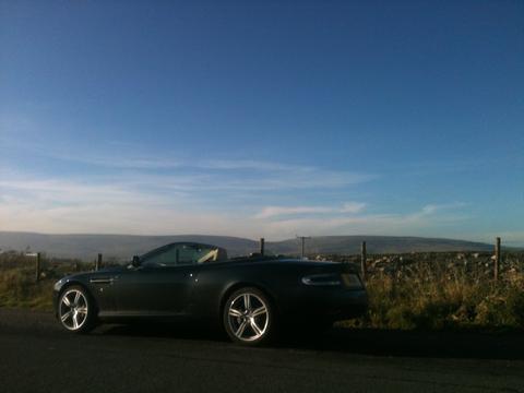 Meteorite, hammerhead, tungsten? - Page 1 - Aston Martin - PistonHeads - The image portrays a sleek black car parked along a road, with a vast, clear blue sky overhead. The car is positioned on the left side of the frame, facing the distant sea in the background. A sturdy stone fence runs next to the road, and a few wooden fence posts are scattered further down the road. The scene suggests a serene and picturesque journey through a coastal landscape.