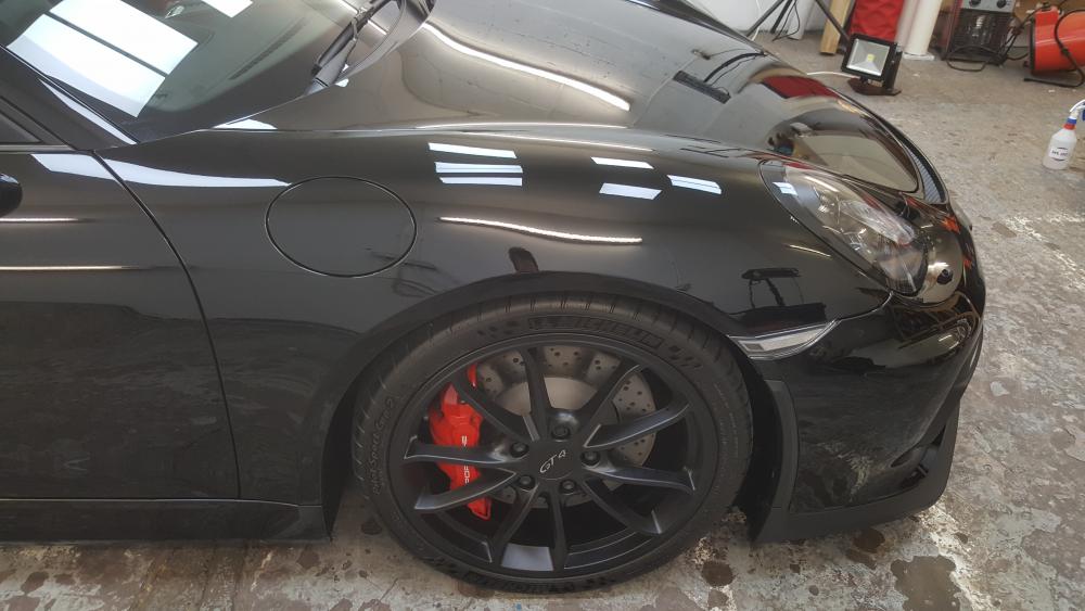 GT4 PPF by Mint condition UK - Page 1 - Porsche General - PistonHeads - The image captures a close-up view of the rear wheel of a black car parked in a repair shop. The reflection on the black car's surface suggests it's a sunny day. Beside the car, a white board can be seen, which is a common feature in such shops for listing repair jobs. Also visible is a red extension cord on the ground, indicating ongoing work or preparations for a work session. The overall scene suggests a professional setting dedicated to vehicle maintenance.