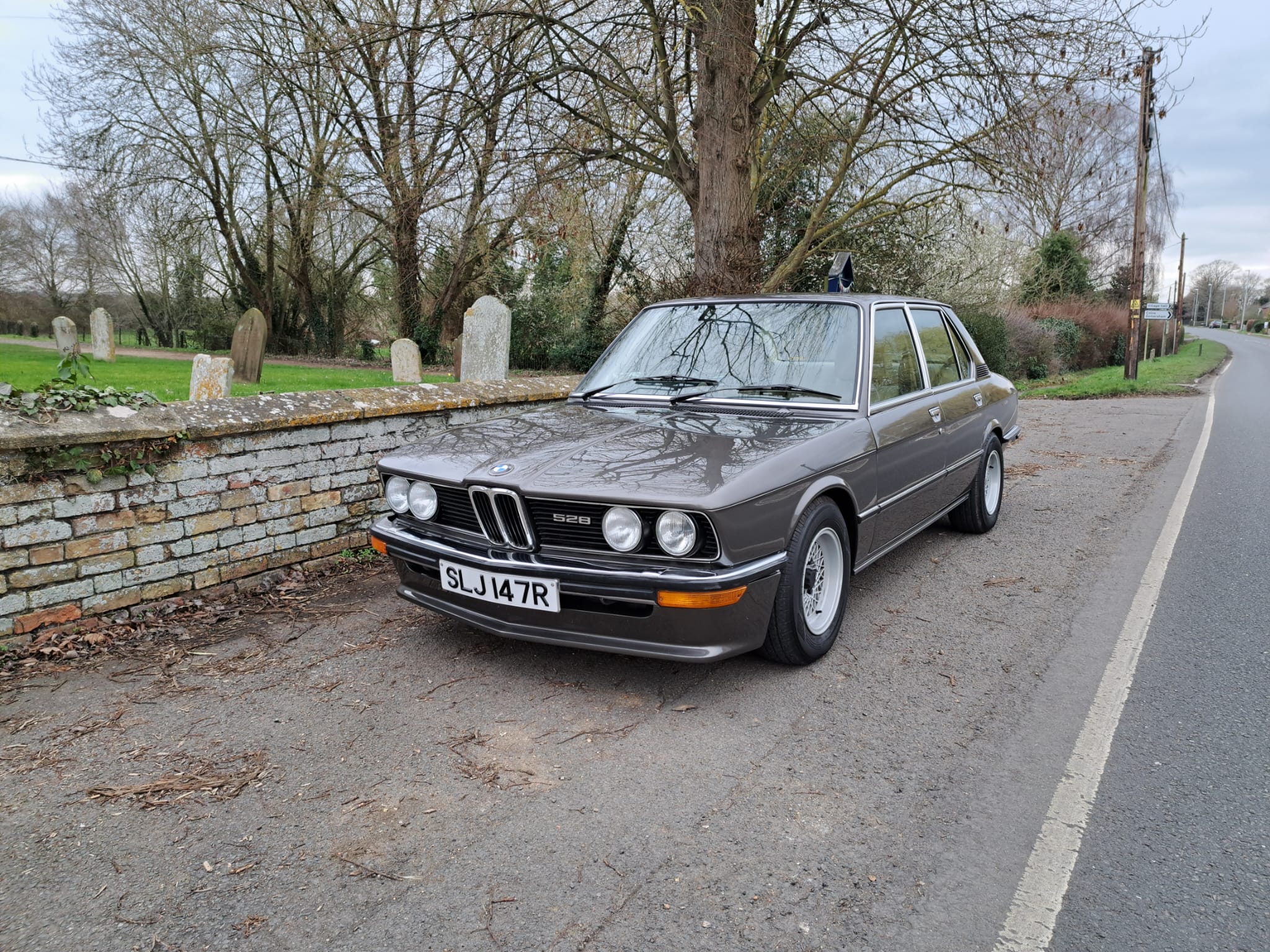 1977 BMW E12 528 - Page 4 - Readers' Cars - PistonHeads UK - The image shows a gray BMW car parked on the side of a road. The car has a license plate that reads "J61 KRJ". In the background, there are trees and a stone wall. The setting appears to be a rural or suburban area, as indicated by the greenery and the presence of a wall. The car's position suggests it may be waiting for someone to return from a nearby location.