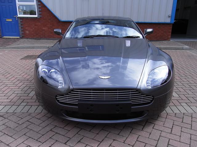 My first Aston - Page 1 - Aston Martin - PistonHeads - The image shows a sleek, gray sports car parked on a brick pavement. The vehicle is facing the camera, and it features a distinctive grille with a prominent logo that is likely associated with the brand. The design of the car is modern and aerodynamic, with a shiny finish that reflects the surroundings. The backdrop includes a faint glimpse of a structure with a blue door, suggesting a location that might be an office or garage.