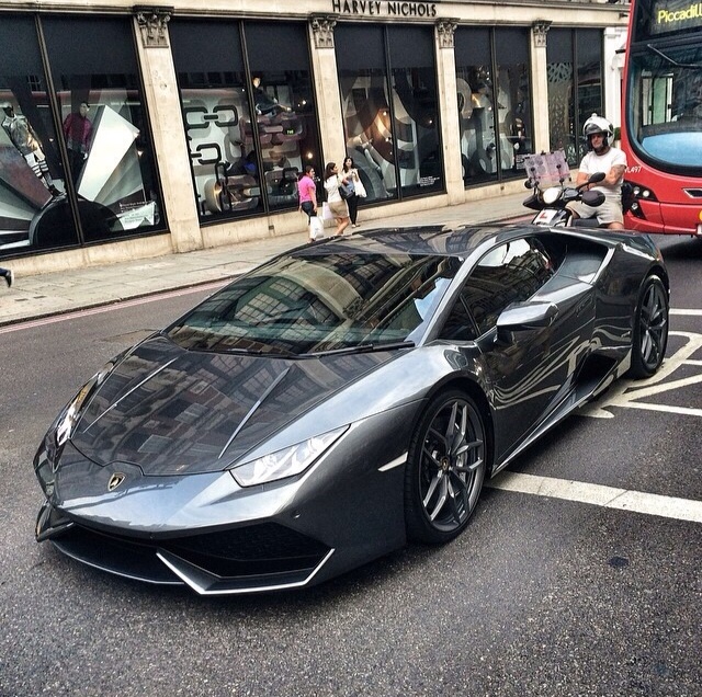 Huracan - Page 1 - Gallardo/Huracan - PistonHeads - The image showcases a scene from an urban setting. Dominating the picture is a sleek, silver sports car with a distinctive design, parked on a city street. The car's shiny exterior reflects off the surrounding environment, adding to its allure. The thin-tire roadster is parked near a bus, indicating a public transport area. Pedestrians can be seen walking by, adding a sense of liveliness to the scene. A large building with a reflective facade forms the backdrop of the image, its massive structure contrasting with the intimate scale of the car.