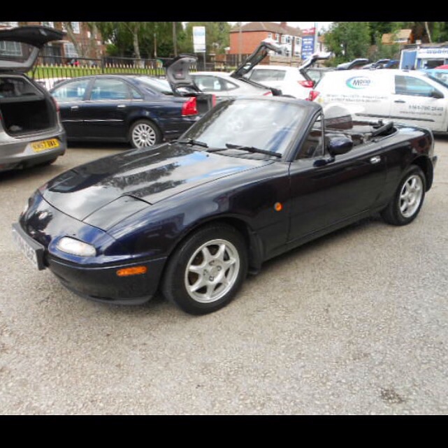 Uh oh I'm going to look at my (potential) 3rd MX5 tonight... - Page 1 - Mazda MX5/Eunos/Miata - PistonHeads - The image shows a black two-door convertible car parked in an empty parking lot. The car is in a rear view angle and appears to be a European model, possibly a BMW Z3 or Z4, given its sleek design and the presence of a soft-top roof. The body of the car is glossy, suggesting it is well-maintained. In the background, there are other cars parked and a white van is visible to the right of the image. The parking lot is contained within a fenced-off space, and an open-box truck can be seen in the far distance. The scene captures a typical day in a parking area.