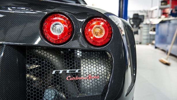 A close up of a red and black motorcycle - Pistonheads - The image captures a close-up view of the back end of a racing car. The primary focus is a pair of prominent red rear lights. Just below the tail lights, there is a red label featuring the word "CARBON." The vehicle's sleek design is accentuated by the smooth black exterior, which contrasts with a noticeable silver visor. In the background, there's a subtle hint of a workshop environment, with metallic structures and equipment visible.